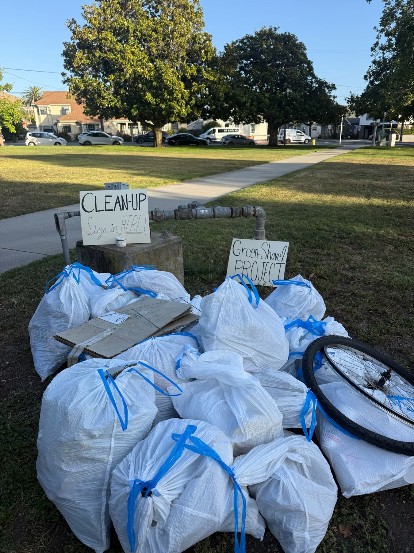 A pile of full trash bags, flattened cardboard and a bike wheel collected at a cleanup, next to Green Shovel Project signs