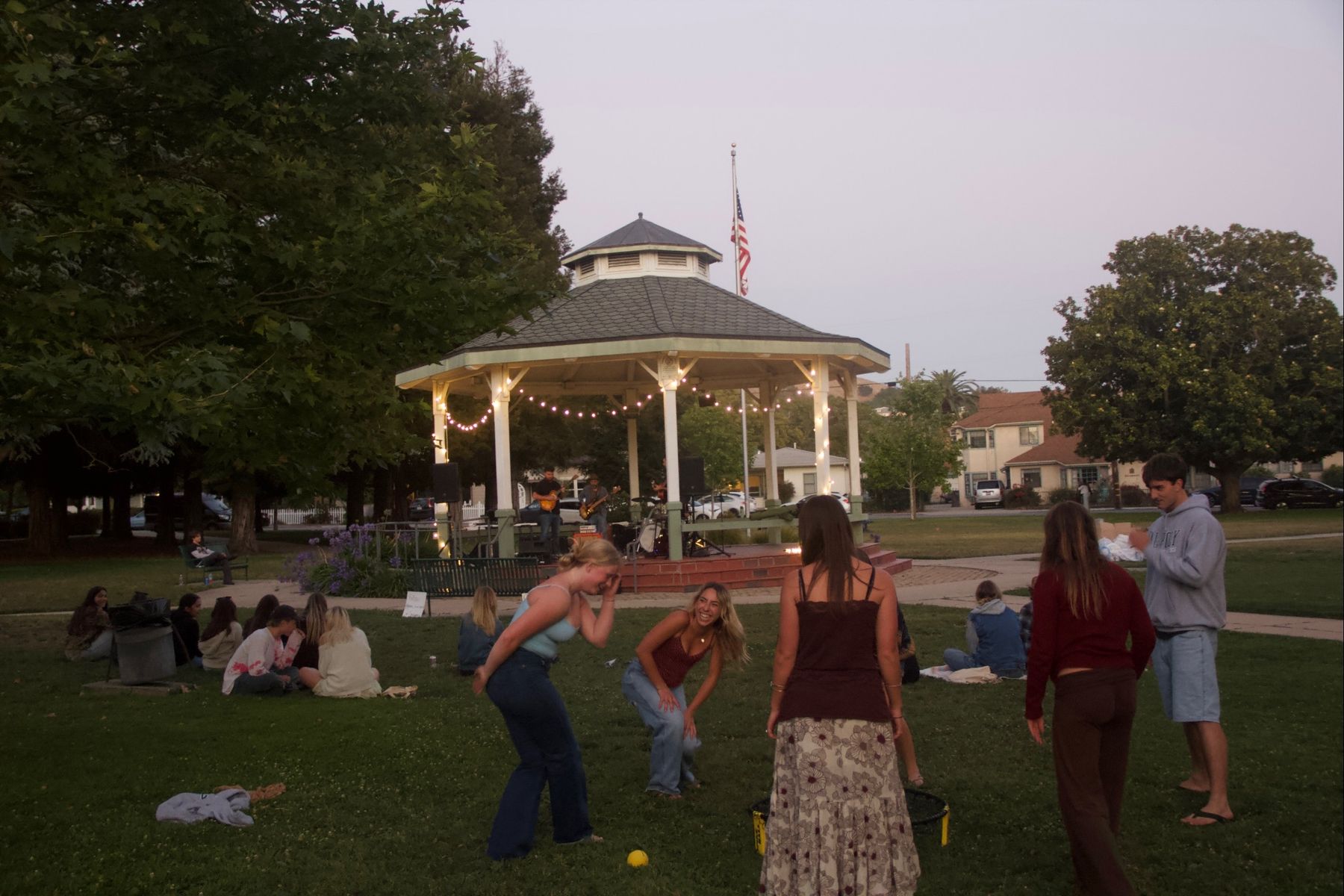 A band playing in a lit gazebo at dusk while students laugh and play spikeball on the lawn
