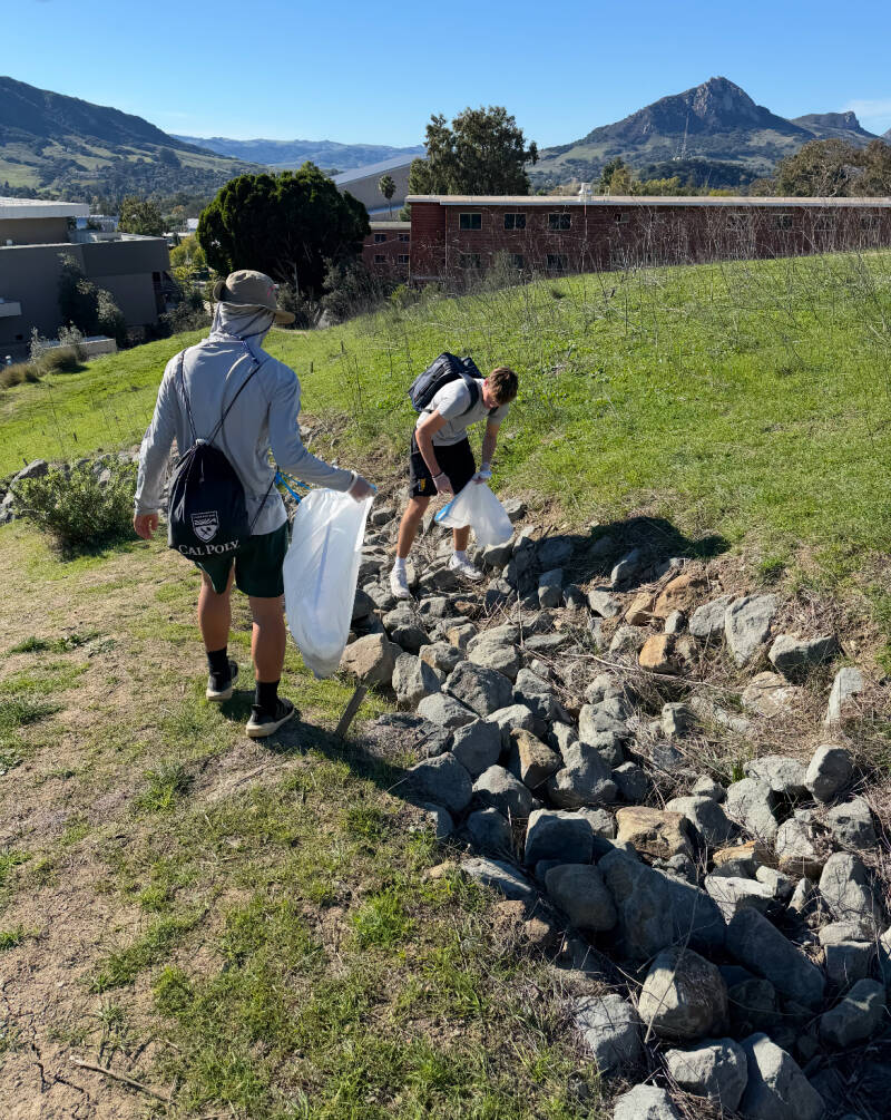 Two volunteers picking up trash along a rocky hillside, with mountains behind them